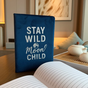 Cuaderno de terciopelo azul oscuro con el mensaje Stay Wild Moon Child captado en un ambiente cálido y moderno.