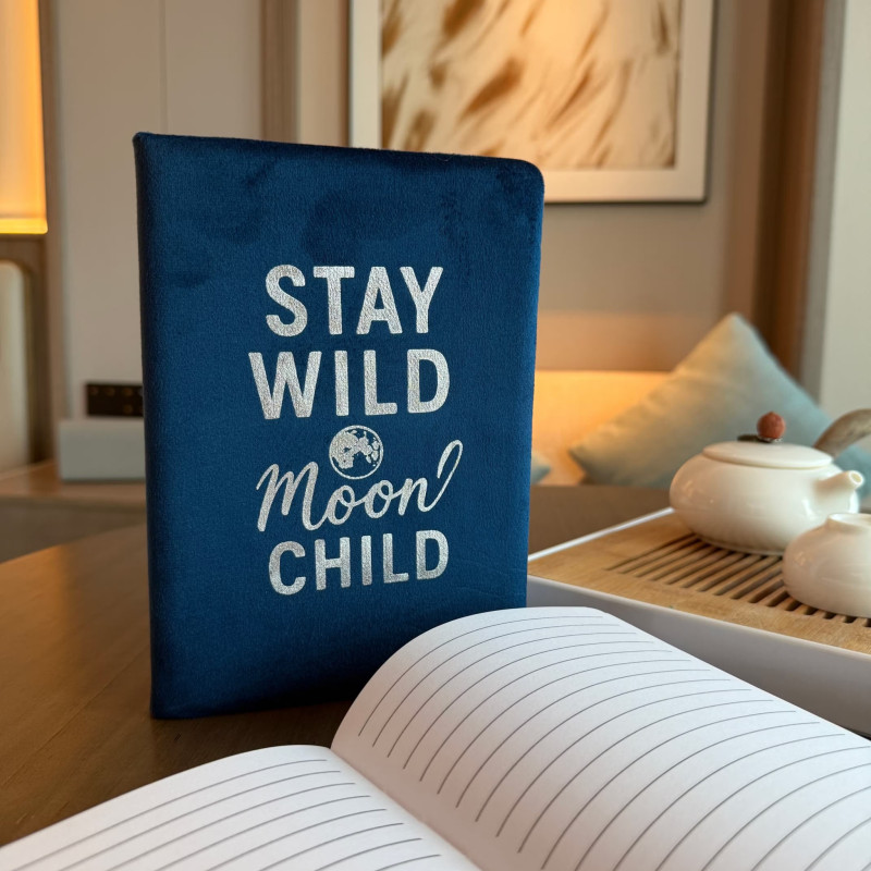 Cuaderno de terciopelo azul oscuro con el mensaje Stay Wild Moon Child captado en un ambiente cálido y moderno.