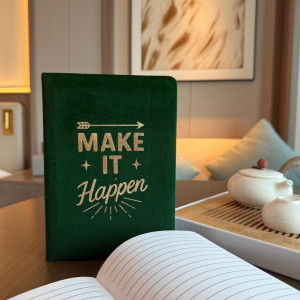 Cuaderno de terciopelo verde oscuro con la frase Haz que suceda, captado en un ambiente cálido e inspirador.